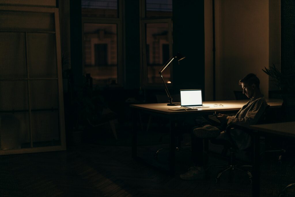 A man sits alone at night in a home study, looking at his phone and laptop, illustrating compulsive pornography use and digital addiction in Singapore.