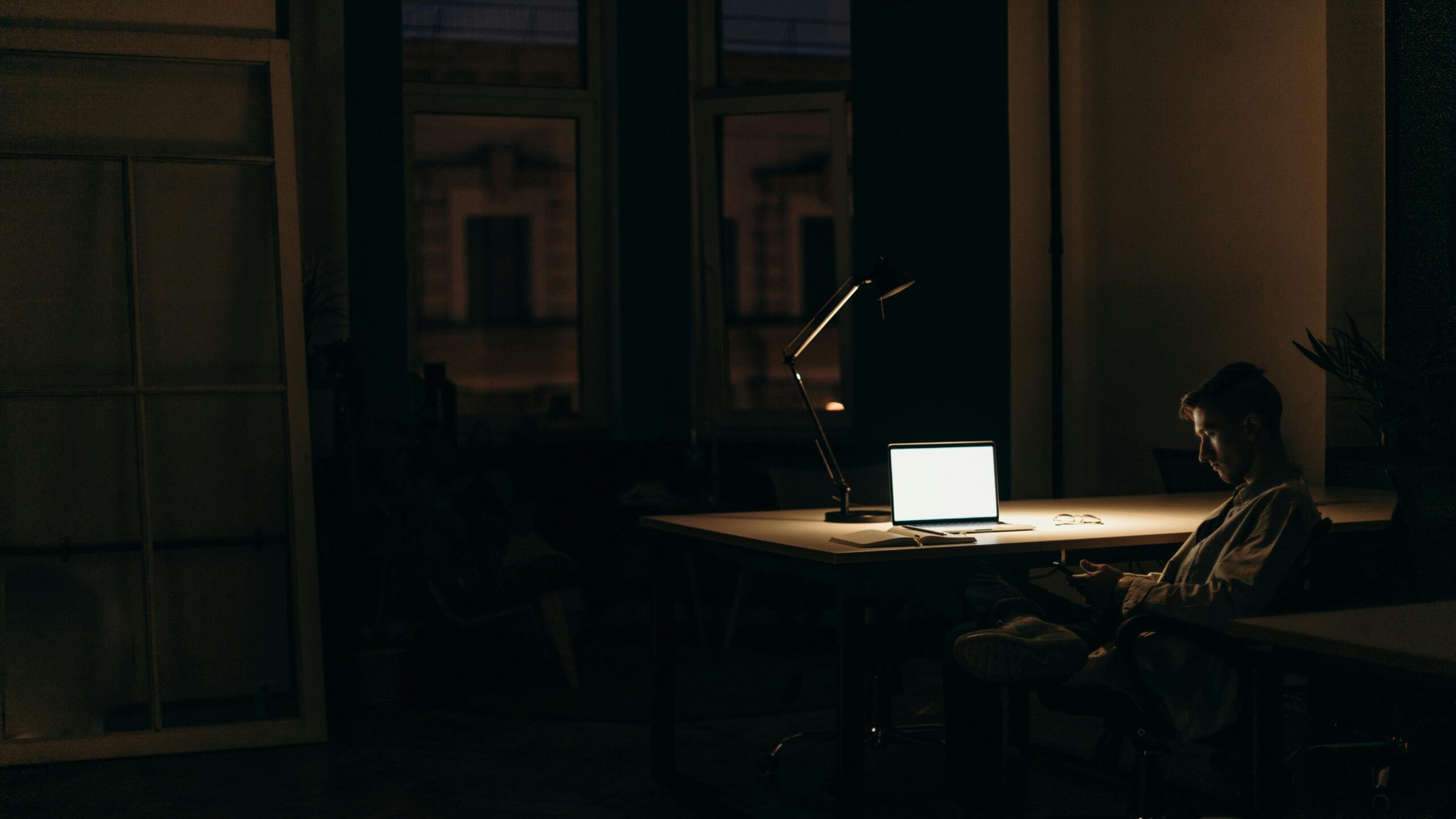 A man sits alone at night in a home study, looking at his phone and laptop, illustrating compulsive pornography use and digital addiction in Singapore.